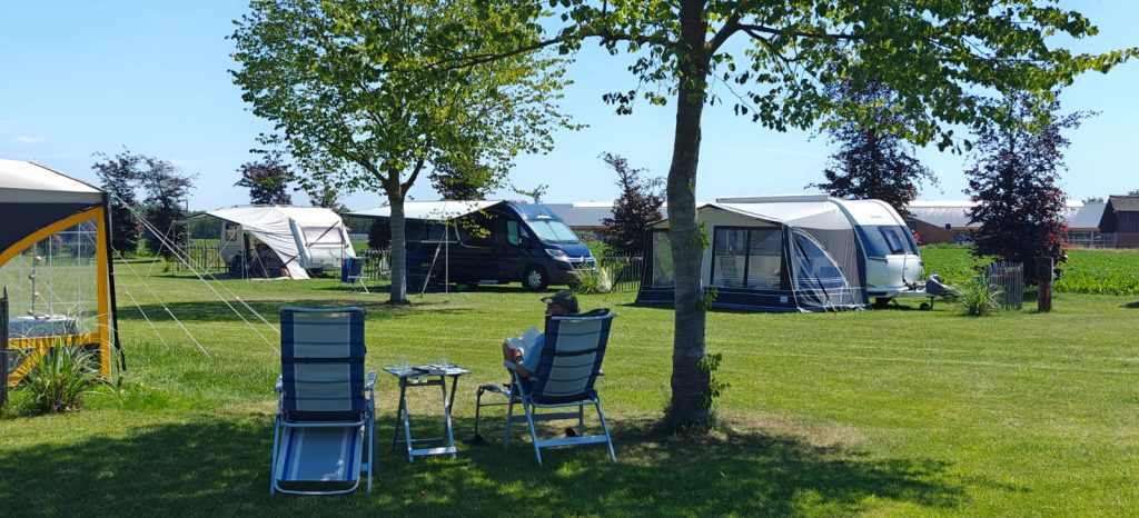 Kampeerveld met caravans en campers op ruime grasplaatsen, met kampeerstoelen in de schaduw van een boom